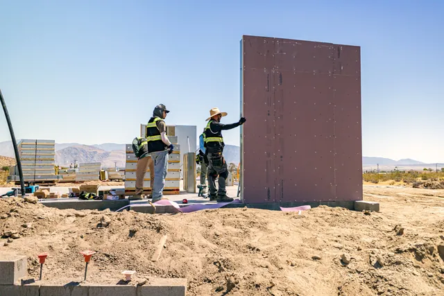 4C crew standing an exterior wall panel by hand on the foundation slab at Borrego Springs, with panel stacks staged behind them